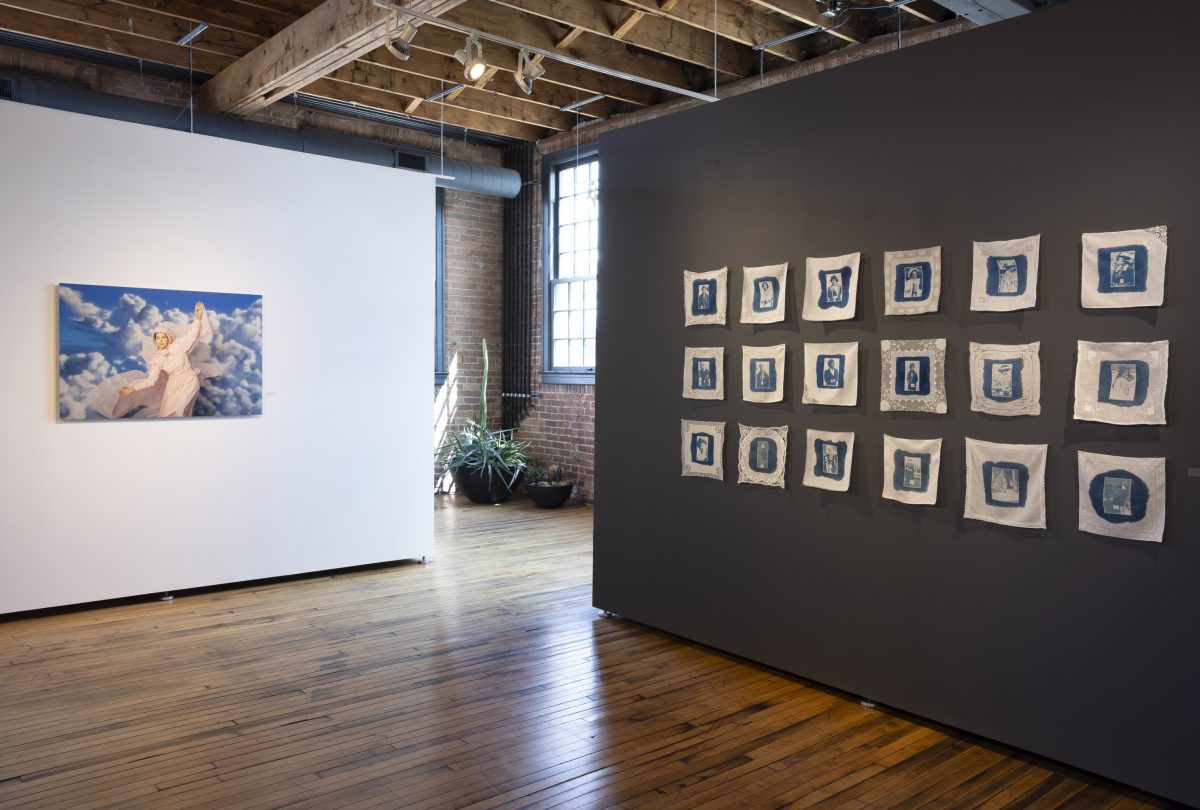 A large color photograph by Angela Ellsworth hangs on a white wall. In it, the artist is dressed in a flowing rose-pink pioneer-style dress with a bonnet and a sheer cape, her arms outstretched as if soaring through a vivid blue sky filled with dramatic white clouds. To the right, on a dark gray wall, 18 vintage white handkerchiefs are arranged in a six-by-three grid. Each handkerchief bears a deep blue cyanotype image of a woman’s portrait—historic mugshots used to track suffragettes. Some handkerchiefs feature lace trim, suggesting domesticity and care.