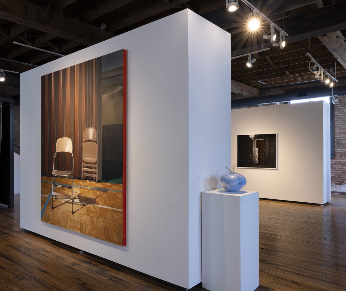 A hyperrealistic painting by Rebecca Campbell depicts two tan metal folding chairs placed on a gymnasium floor with parquet wood patterning. The chairs are set against a backdrop of tall, dark wood-paneled folding doors, evoking the interior of an LDS church building. One chair is upright and confronts the viewer; the other is folded and leans against the wall. In front of the painting is a light blue ceramic vessel with a curved wooden handle, placed on a white pedestal. In the background, a black-and-white photograph of a metal gate is visible, echoing the visual motifs of closure and concealment.
