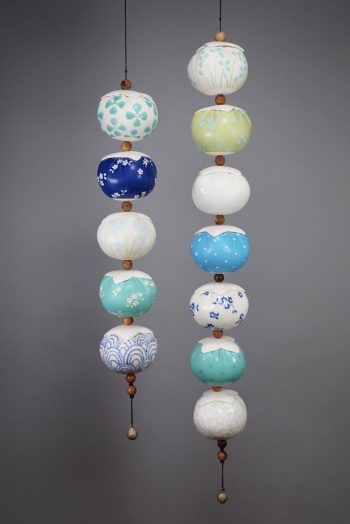 Two vertical strands of spherical ceramic forms hang side by side against a gray background. Each ceramic bead is shaped like a soft pincushion and decorated with intricate patterns in blue, white, green, and yellow glazes, including polka dots, floral motifs, chevrons, and scallop designs. The beads are spaced with round wooden separators and suspended from black cords. Each strand ends with a wooden bead and a small brass bell. The overall effect is delicate and rhythmic, evoking handcrafts and heirloom sewing traditions.