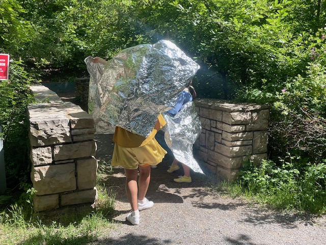 Two dancers wearing metallic reflective fabric raise it overhead like wings near a stone entryway in a forested area.