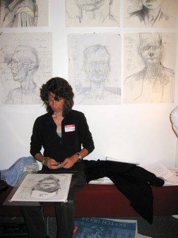A woman seated and sketching a portrait, surrounded by expressive pencil drawings of faces pinned to the wall behind her.