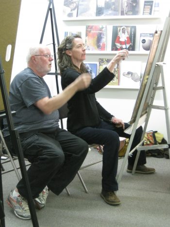 A woman and a man seated side by side at easels, mid-discussion, with art books and paintings displayed on the wall behind them.