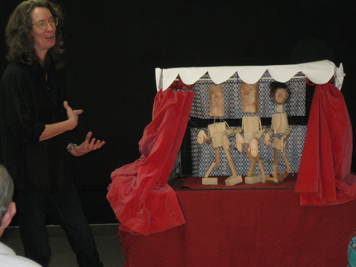 A woman presenting a puppet performance, gesturing beside a red miniature stage with three wooden marionettes.