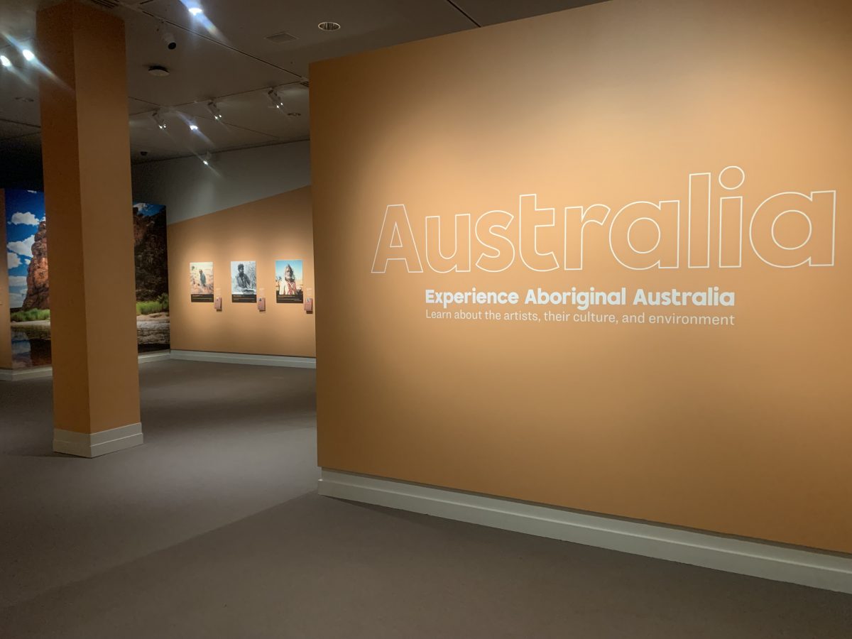 Exhibition wall with large text reading “Australia: Experience Aboriginal Australia,” surrounded by images and orange-toned walls.