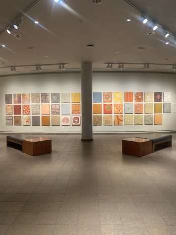 Gallery installation of square-format Aboriginal paintings arranged in a large grid on a white wall, with benches in the foreground.