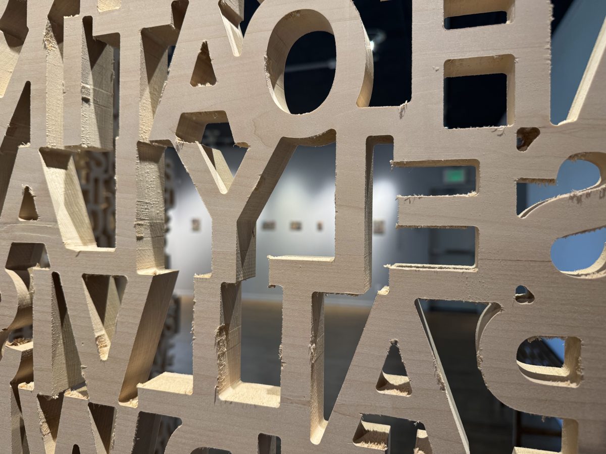 Close-up of wooden text cutouts, focusing on the letter shapes with blurred collage artworks visible in the background.