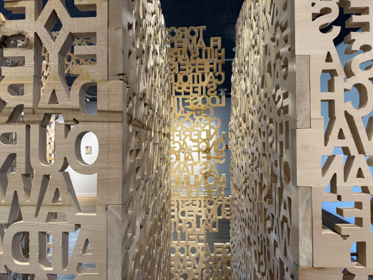 Close-up view of intricately carved wooden panels with cut-out letters forming dense walls of text, part of a large-scale installation in a gallery space.