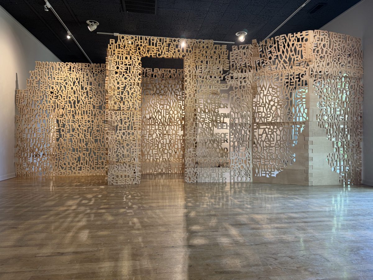 Wide-angle view of an expansive wooden installation composed of interconnected panels with cut-out text casting shadows on the gallery floor.