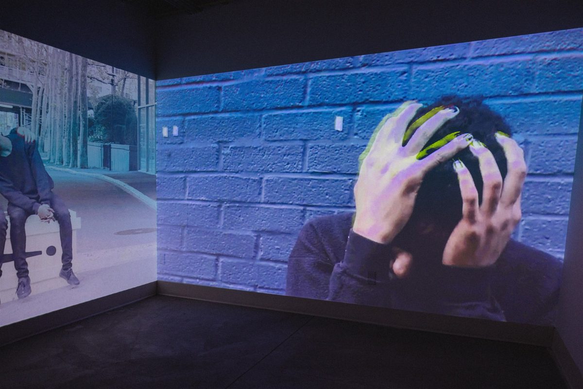 Large projected video shows a close-up of a person with hands gripping their head against a blue brick wall, with another figure seated to the side.