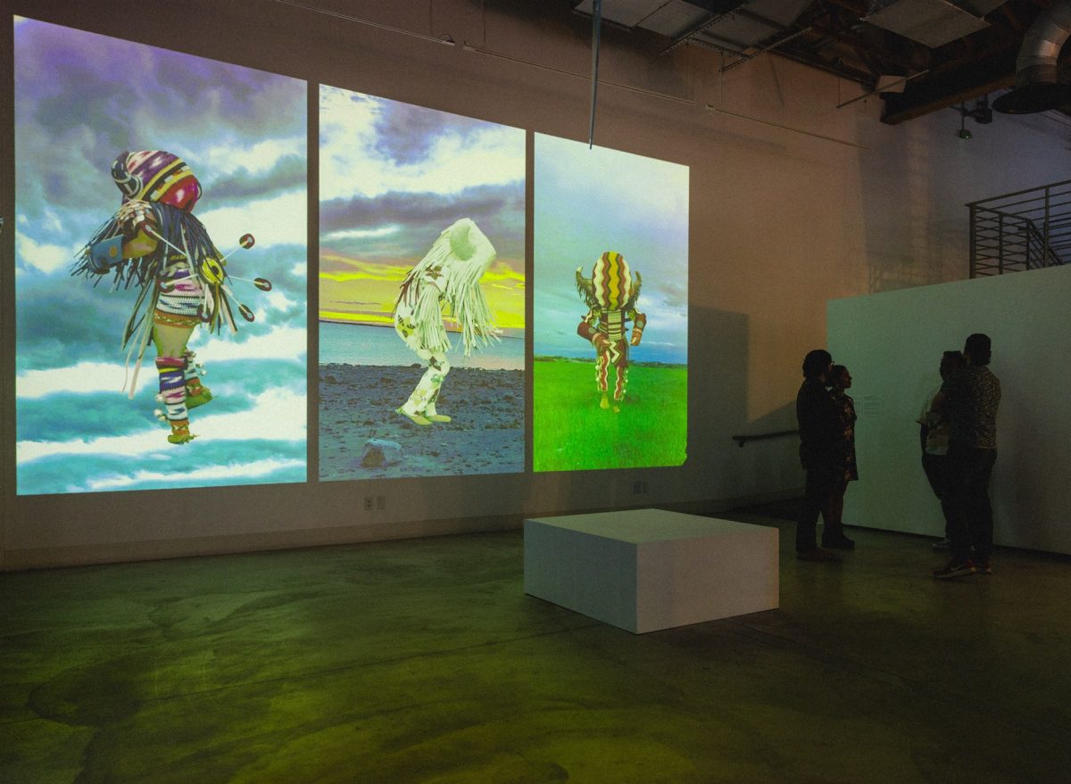 Three large video projections on a gallery wall show figures in colorful buffalo regalia dancing against backdrops of sky, sea, and grassland, with silhouetted viewers standing nearby.