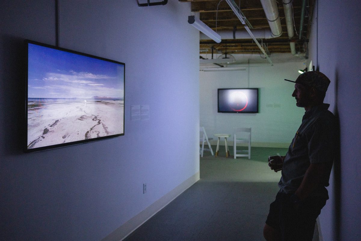 A man leans against a gallery wall watching a video screen that shows a desolate landscape with wide, cracked earth under a blue sky; another video glows faintly in the background.