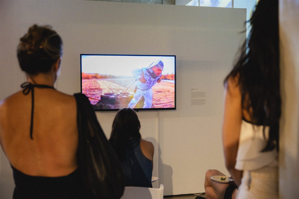 People view a wall-mounted screen showing a video of an artist in white clothing pulling tires across sandy soil in the desert.