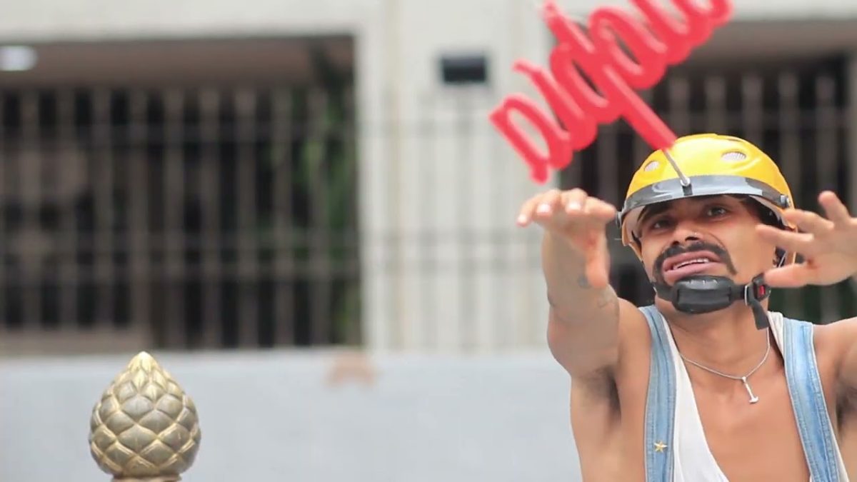 A man wearing a yellow helmet and overalls reaches toward a red sculpture of the word “utopia,” performing outdoors in front of a stone building.