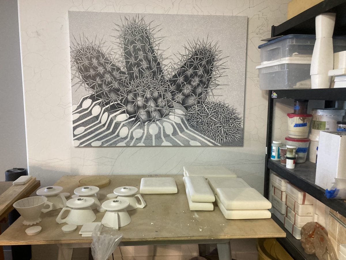 A studio worktable with ceramic molds and bisque forms arranged below a large black-and-white drawing of cacti intertwined with industrial piping and circuitry.