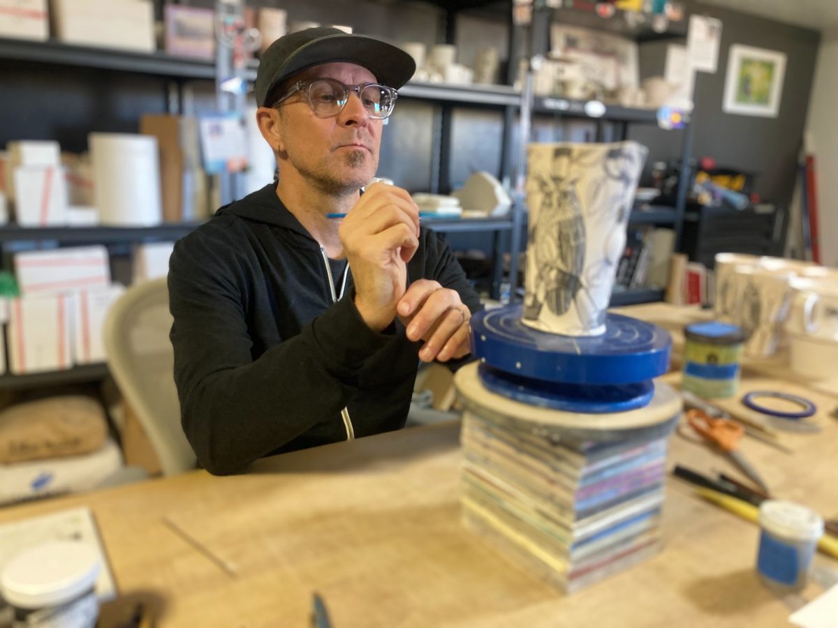 Artist Jason Walker, wearing glasses and a cap, concentrates as he paints detailed imagery onto a ceramic vessel on a rotating stand in his studio.