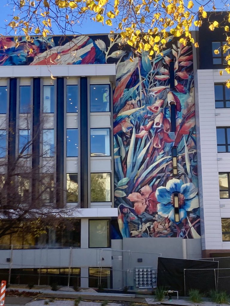 A tall, multi-story mural by Chuck Berrett covering the side of a renovated building. The artwork features large, stylized botanical forms in blues, reds, and warm tones, weaving between dark vertical window columns. Yellow autumn leaves frame the top of the image.