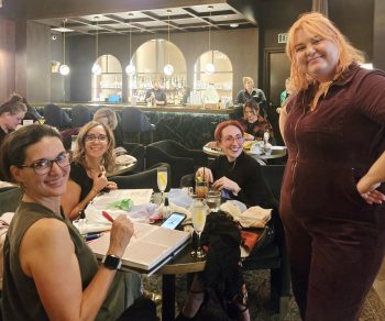 People gathered at small tables inside Edison House, participating in an art activity; a group of women smile toward the camera as they work on creative projects in the club’s lounge.