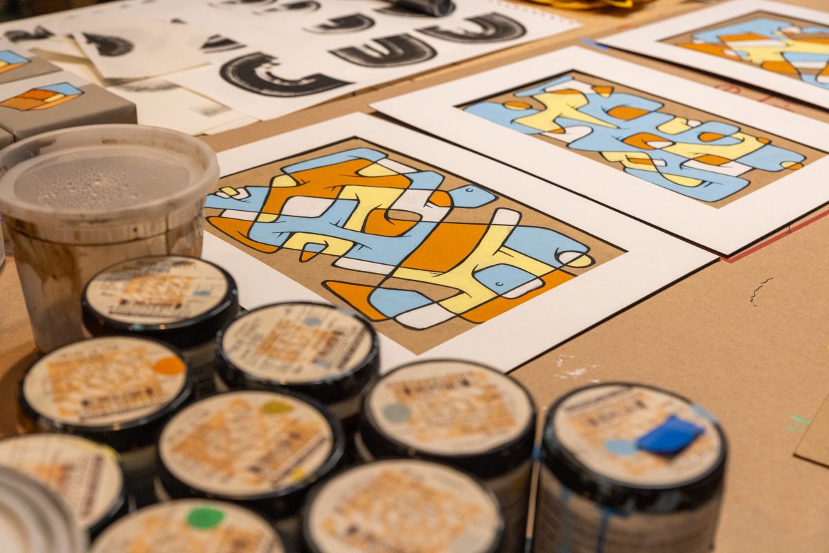 A table in Ryan Harrington’s studio covered with acrylic paints, sketch papers, and colorful block-head character illustrations in yellow, blue, and orange.