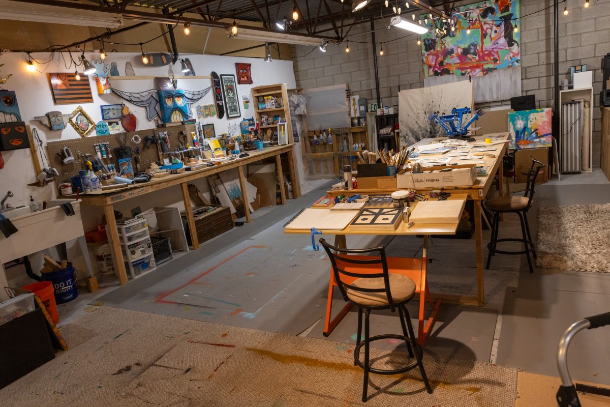 Wide view of Ryan Harrington’s Midvale studio, featuring workbenches, tools, paintings, and an arrangement of tables covered with sketches and art supplies.