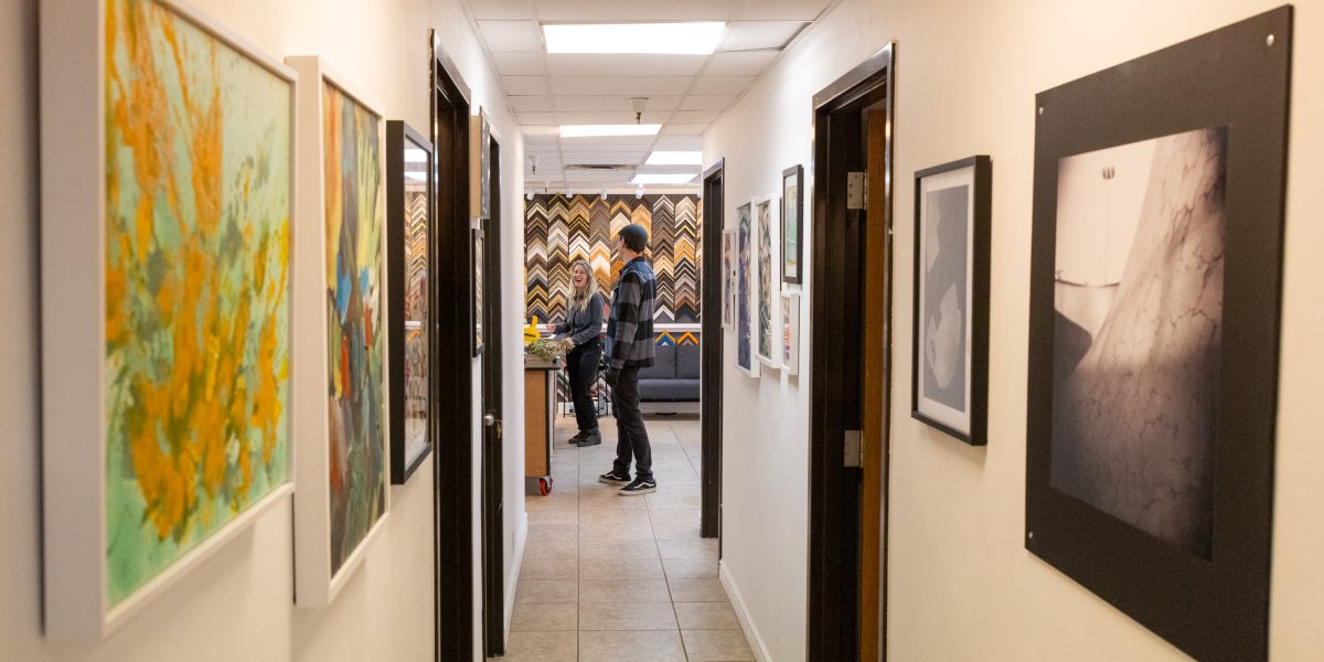 A hallway at Harrington Art Studio lined with framed artworks on both sides, leading to a framing area where two people talk near a wall of colorful frame samples.