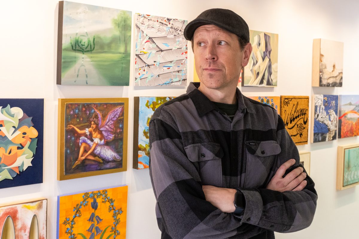 Artist Ryan Harrington standing with folded arms in his gallery space, surrounded by a grid of diverse artworks by Utah artists.