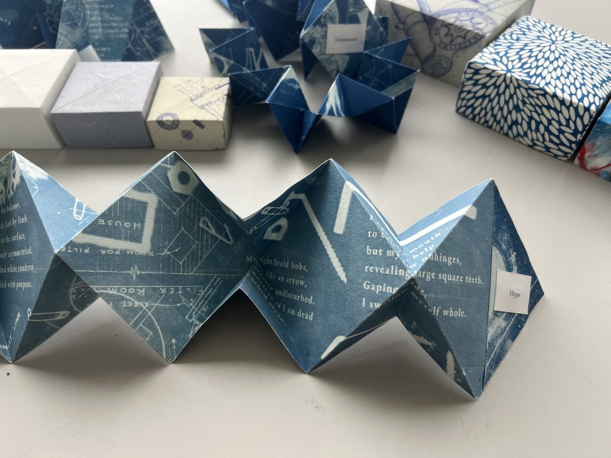 A blue cyanotype artist book arranged in a zigzag fold on a tabletop, its pages printed with layered text and blueprint-like imagery; additional folded book objects and patterned boxes sit blurred in the background.