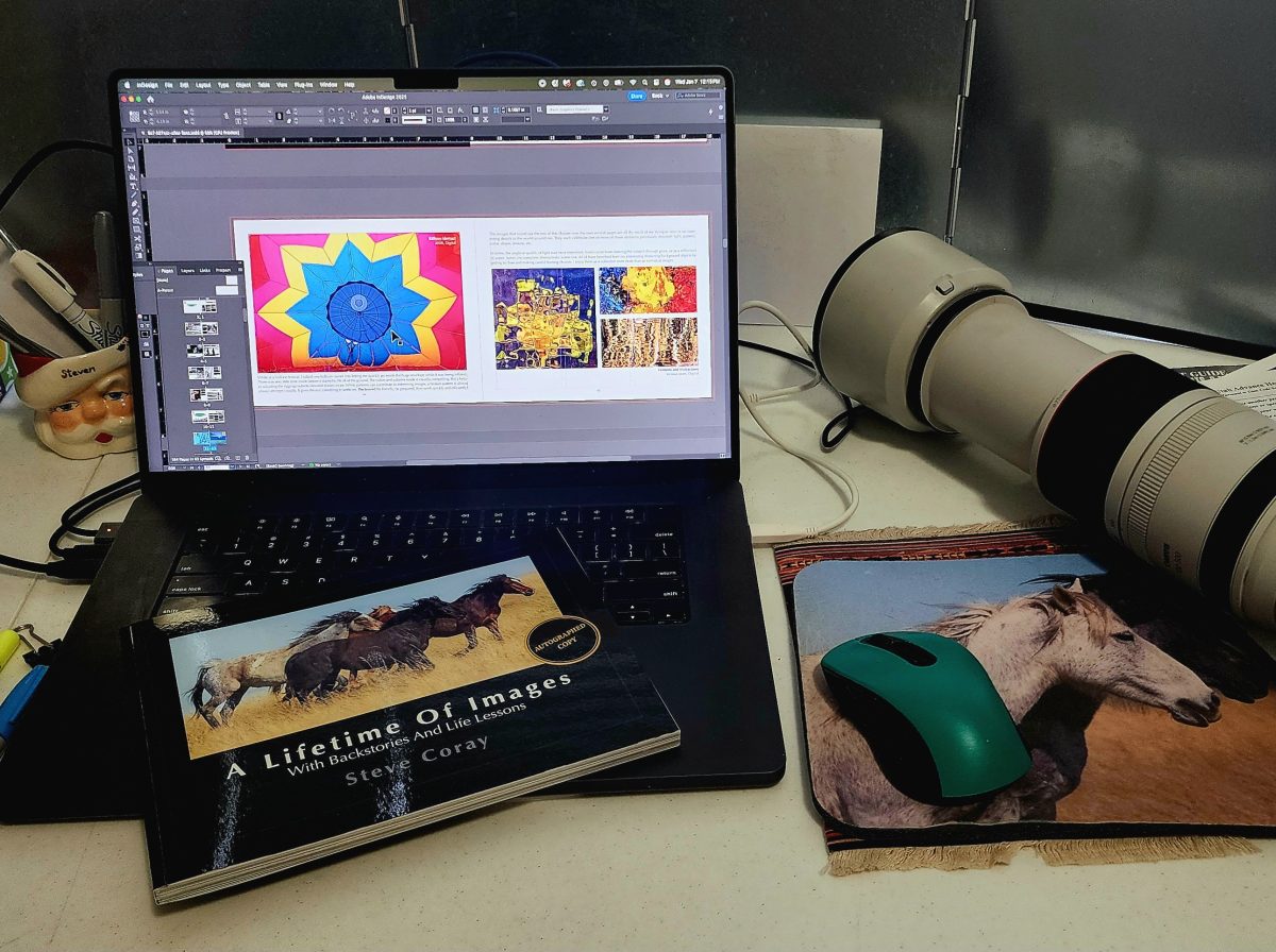 Desk scene with an open laptop showing a book layout program, a large telephoto camera lens, and a printed copy of A Lifetime of Images resting on the keyboard.