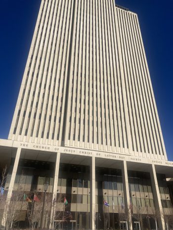 Low-angle view of the LDS Church Office Building tower in Salt Lake City, a tall modernist high-rise with narrow vertical window bands against a deep blue sky.