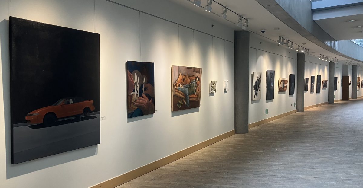 Wide view of a long gallery hallway with paintings installed along a curved white wall, supported by concrete columns and illuminated by ceiling track lights.