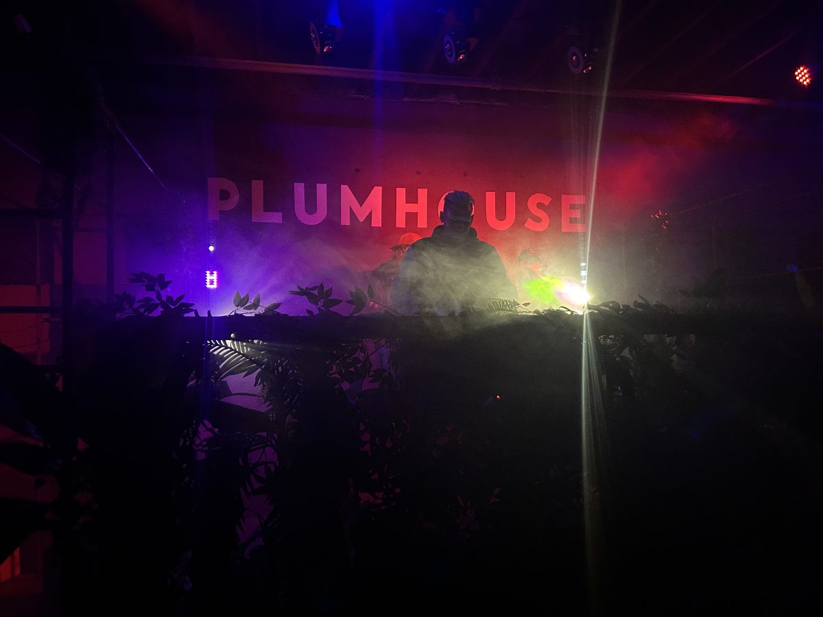DJ silhouetted behind a booth at Plumhouse, with colorful stage lights, haze, and the word “PLUMHOUSE” visible on the back wall.