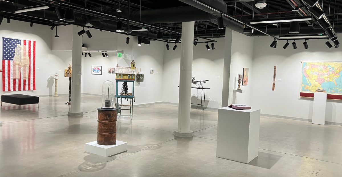 Wide view of a contemporary gallery space with sculptures, altered books, and a large American flag artwork on the wall, arranged across an open floor with white pedestals.