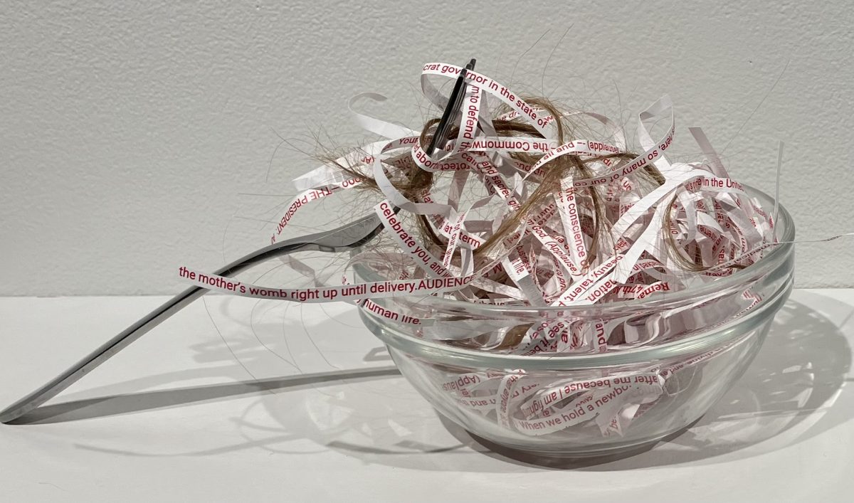 Glass bowl filled with shredded strips of printed text mixed with strands of hair, with metal utensils placed beside it.