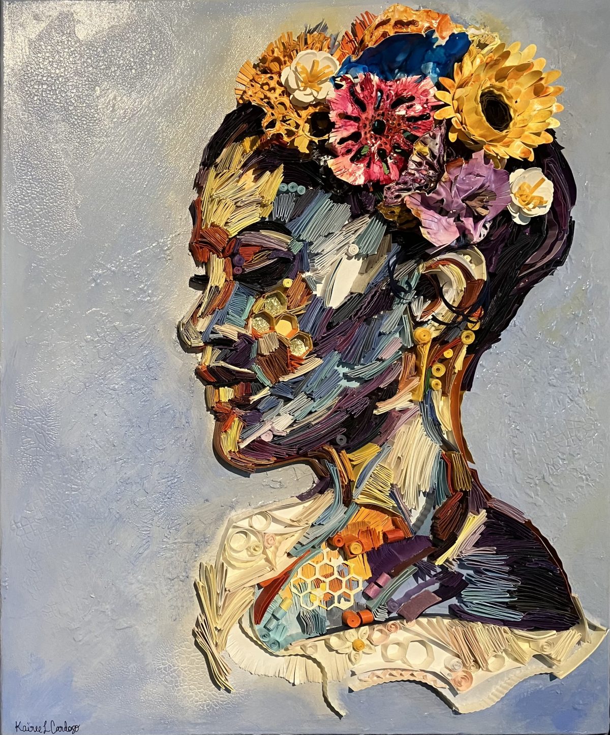 A textured, three-dimensional portrait by Kairee Cardoso titled 'Floral Harmony.' The side profile of a woman’s face is created using colorful relief materials resembling quilled paper. Her head is adorned with vibrant flowers, adding dimension and intricate detailing.