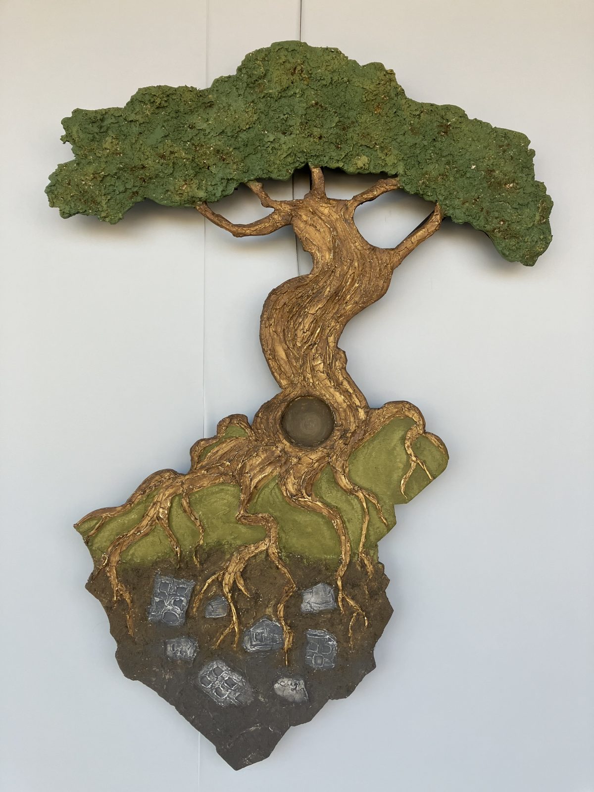 A wall sculpture depicting a stylized tree with twisting roots extending into the earth, anchoring it to a rocky foundation. The textured foliage contrasts with the smooth, golden roots that seem to grasp at embedded stone shapes.