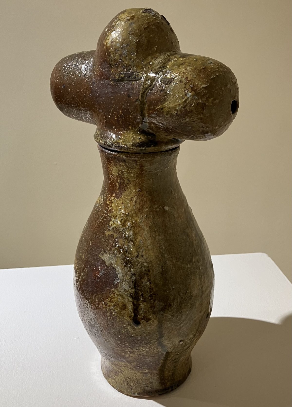 A unique ceramic sculpture with a cross-shaped top and an organic, earthy glaze, standing on a narrow, curved base.