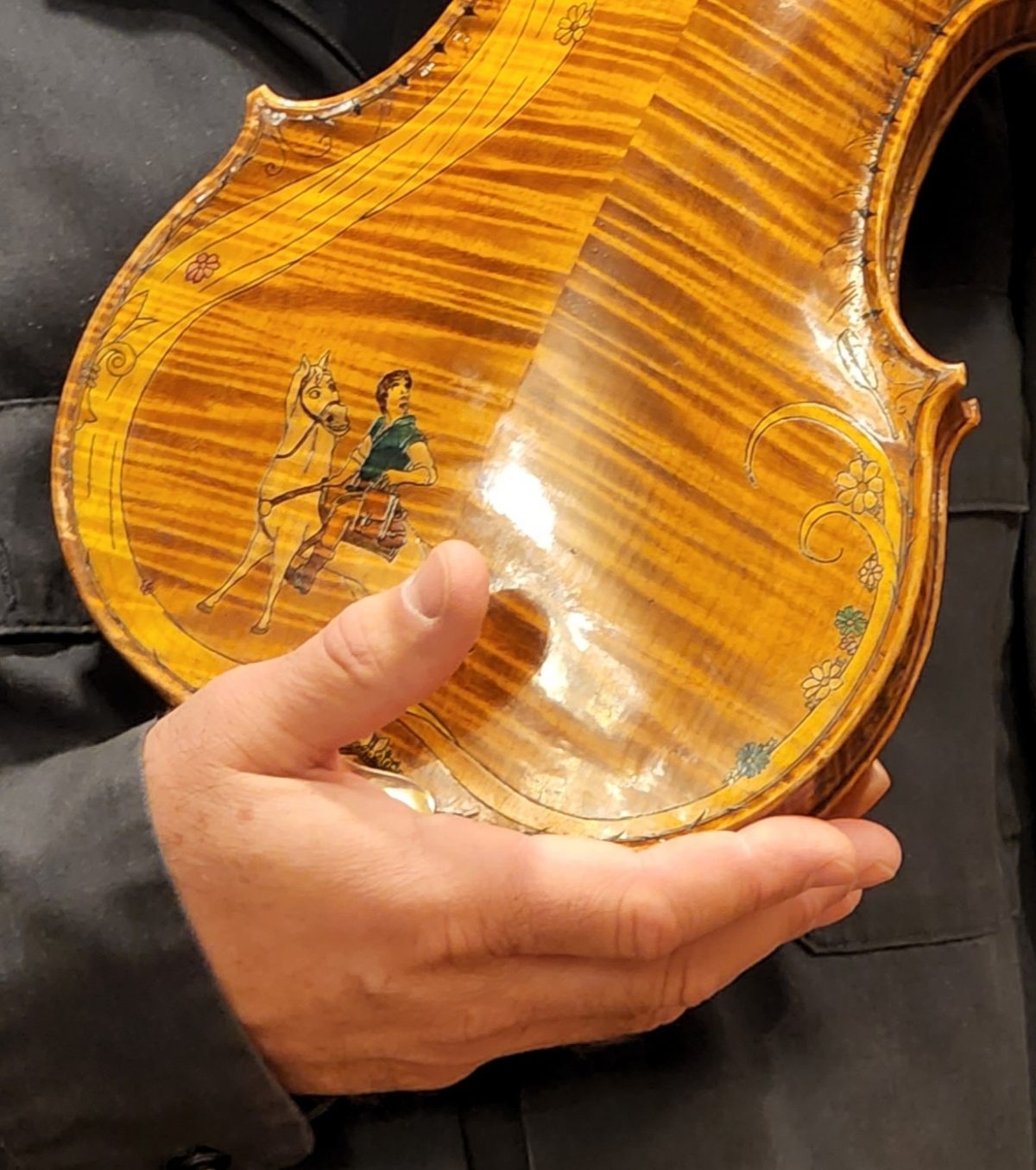 violin back plate with a richly figured maple pattern, decorated with painted illustrations of a person with a horse and floral embellishments, held by a person.