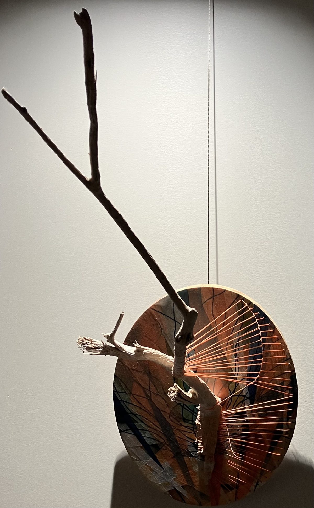 A mixed-media sculpture featuring a wooden branch extending outward from a circular base. The base is painted with abstract tree-like patterns in earthy tones and is adorned with orange string radiating outward in a fan-like arrangement, creating a dynamic interplay between natural and geometric elements.