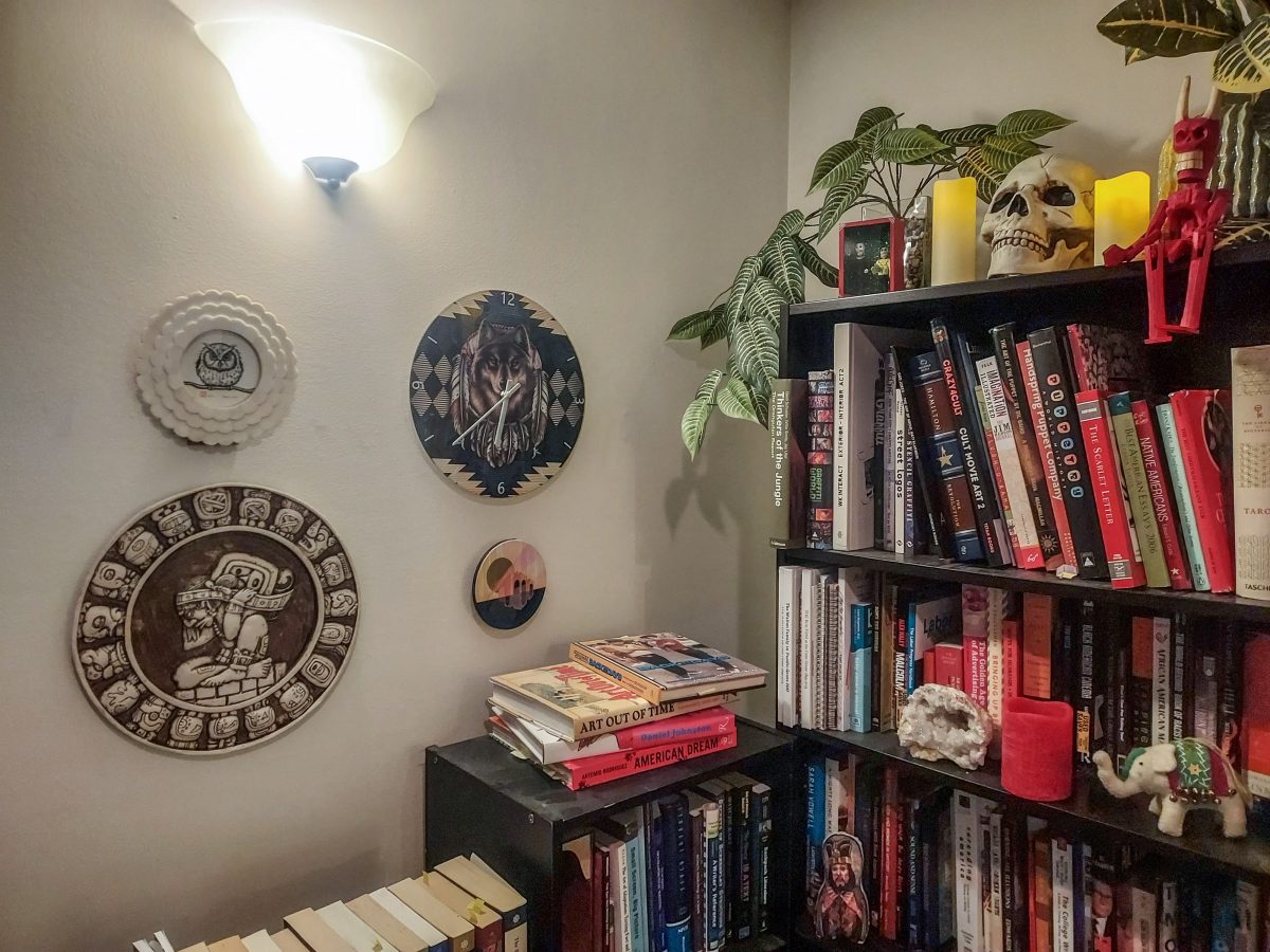 A bookshelf filled with books and decorative items, including a ceramic skull, plants, and wall-mounted art pieces, such as a clock and a detailed ceramic plate.
