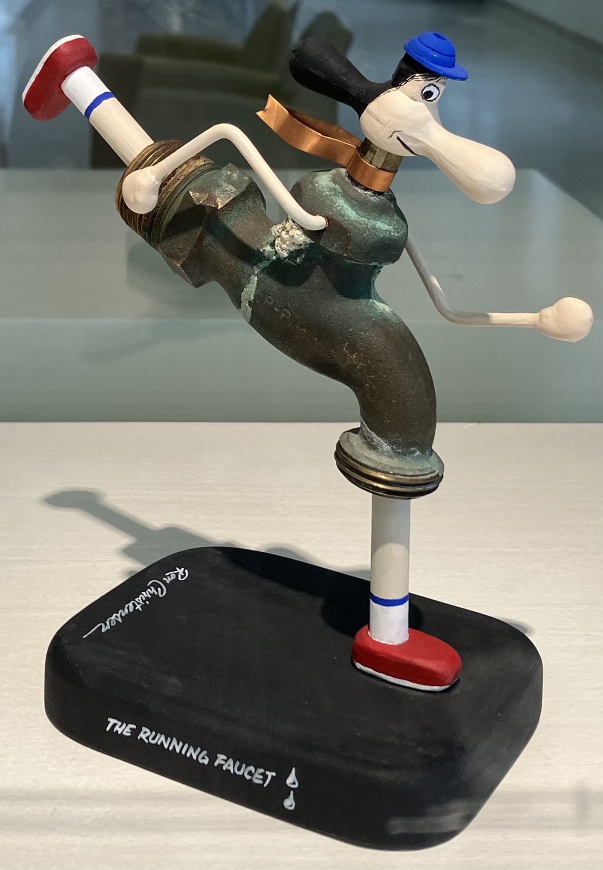 A whimsical sculpture titled "The Running Faucet" by Ron Christensen, depicting a faucet anthropomorphized as a running figure, complete with a blue cap, red shoes, and dynamic posture.