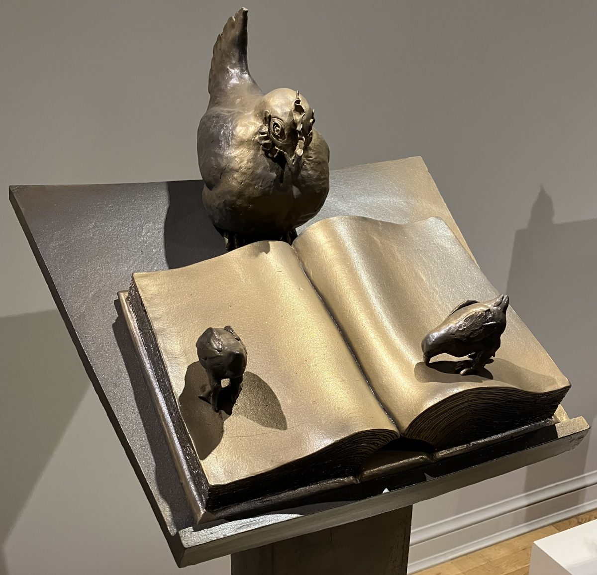 A metallic sculpture of a chicken standing over an open book, accompanied by two small bird-like figures peering at the pages. The bronze-colored piece is positioned on a raised platform, combining literary and organic themes.
