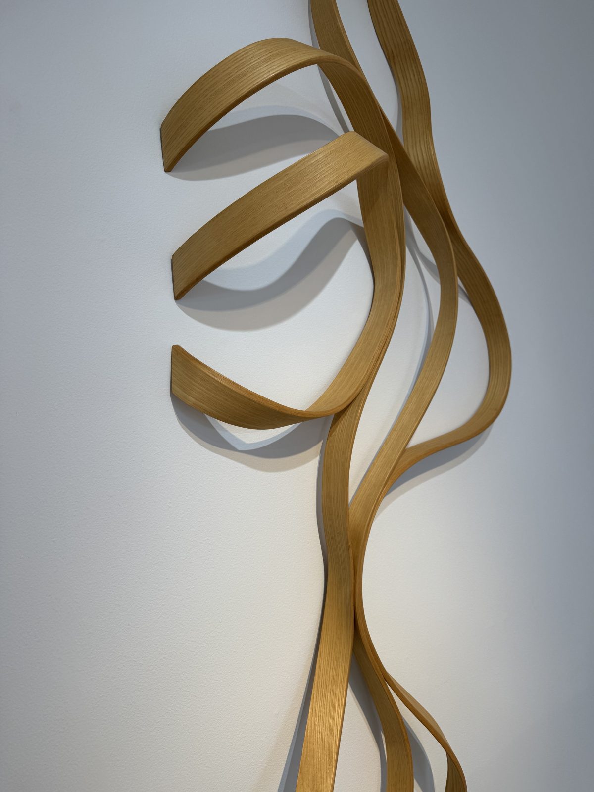 A delicate wooden wall sculpture in golden tones with flowing curves, resembling swirling flames or tendrils of smoke. The piece casts gentle shadows on the white wall behind it.