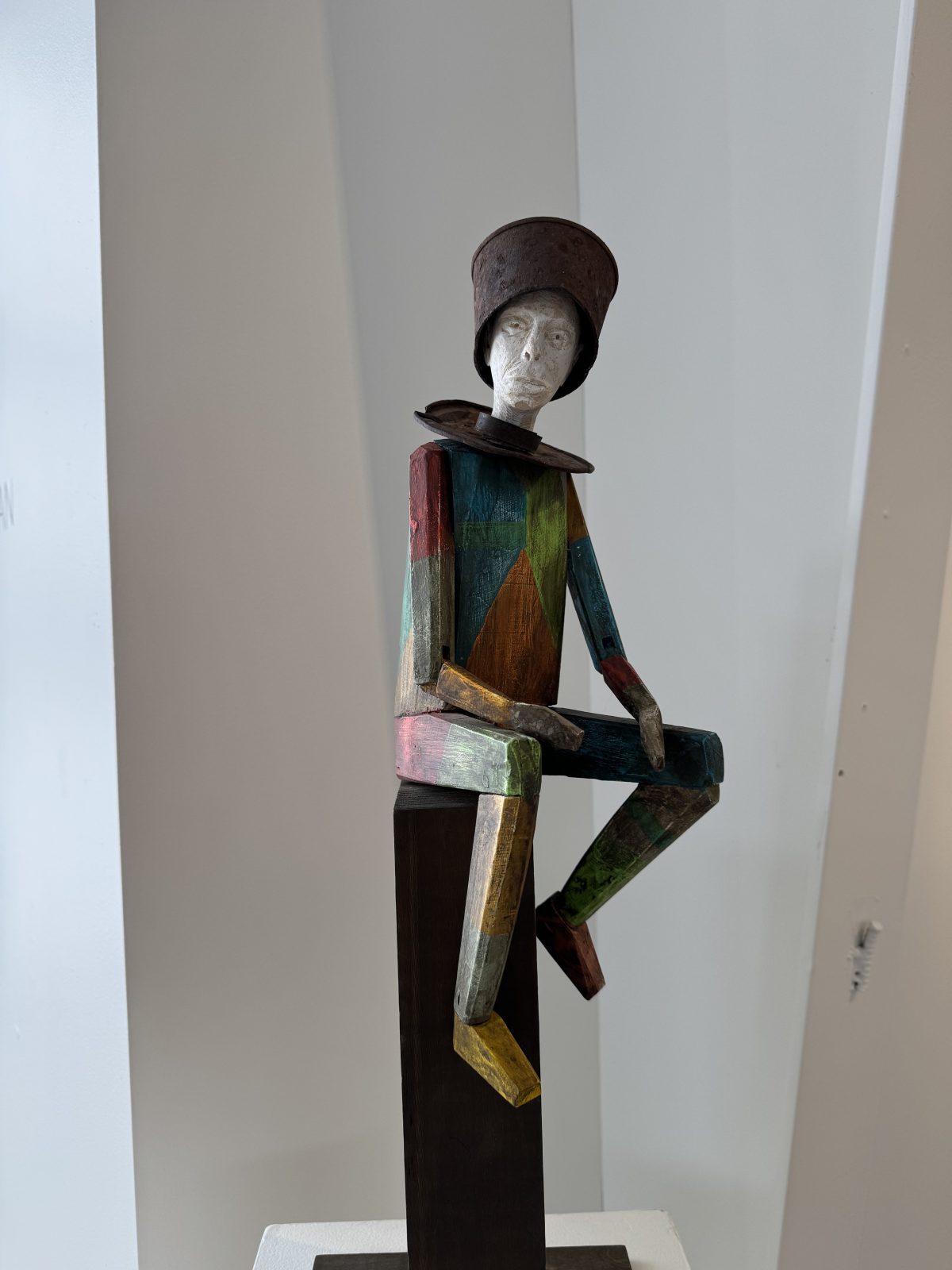 A seated wooden sculpture with a painted patchwork body and a rusted metal bucket hat. The figure’s abstract, geometric limbs and contemplative pose give it a whimsical yet reflective character.