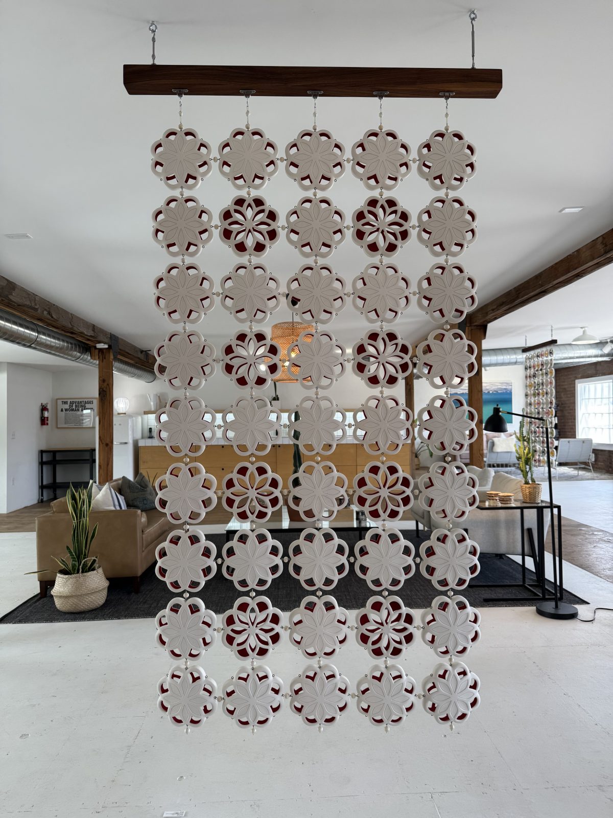 Hanging panel of white, floral-patterned circular ceramic pieces arranged in rows, suspended in front of a modern lounge area with natural light and wooden beams.