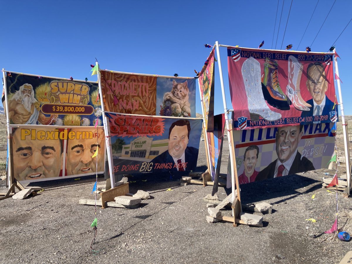 A series of hand-painted banners in a desert setting, depicting spoof advertisements including “Super Win,” “Spaghetti Man,” and “National Debt Relief,” arranged on a structure held up with concrete blocks.