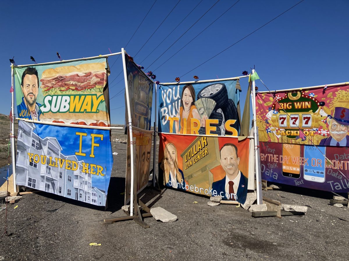 Outdoor installation of brightly colored hand-painted banners referencing American advertising, including parodies of Subway, real estate, tire sales, and payday loans, set against a clear blue sky.