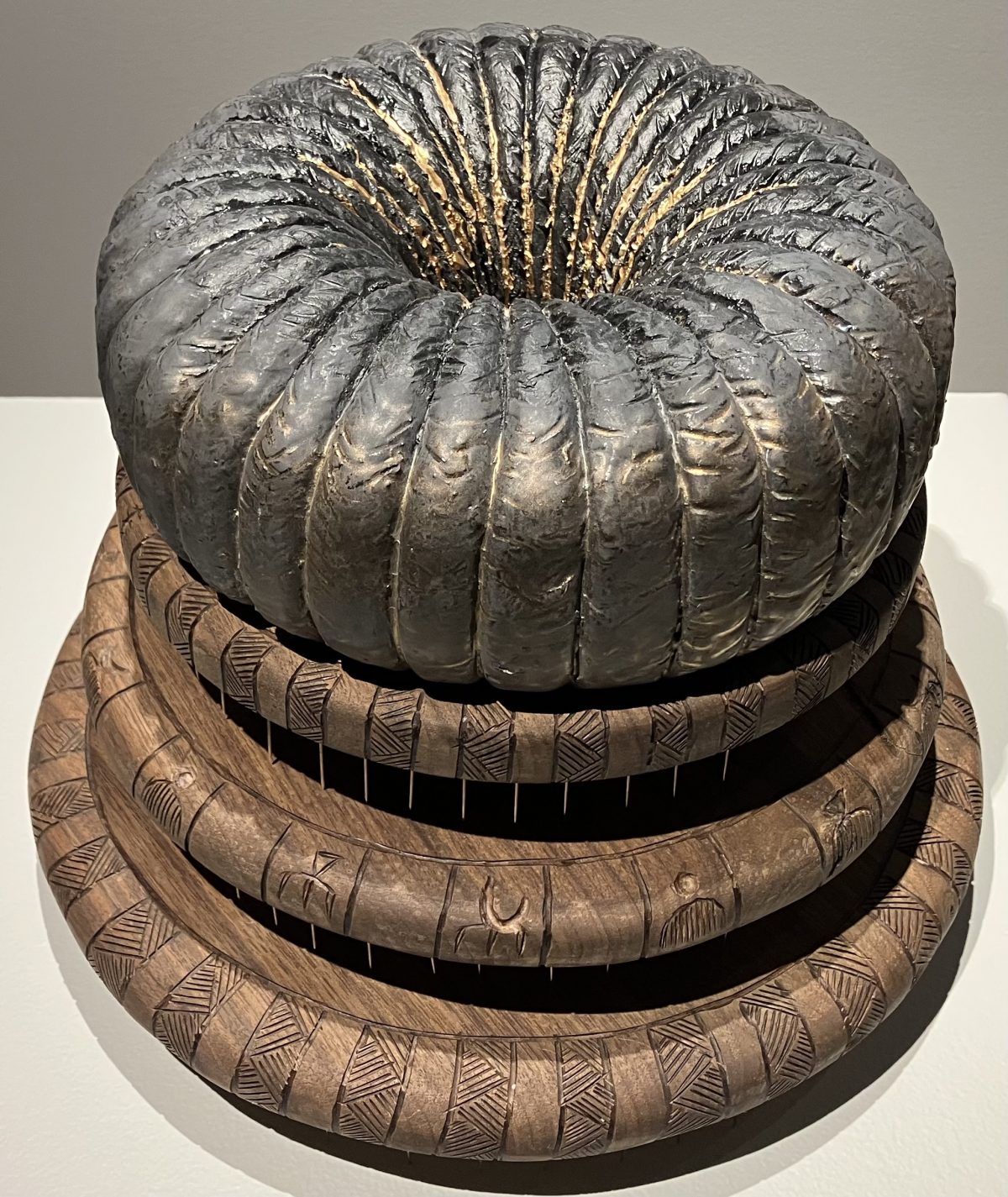 A sculptural piece featuring a large, circular ceramic form with intricate patterns, stacked on engraved wooden rings. The dark, metallic top has a textured surface, while the wooden layers are adorned with symbolic carvings.