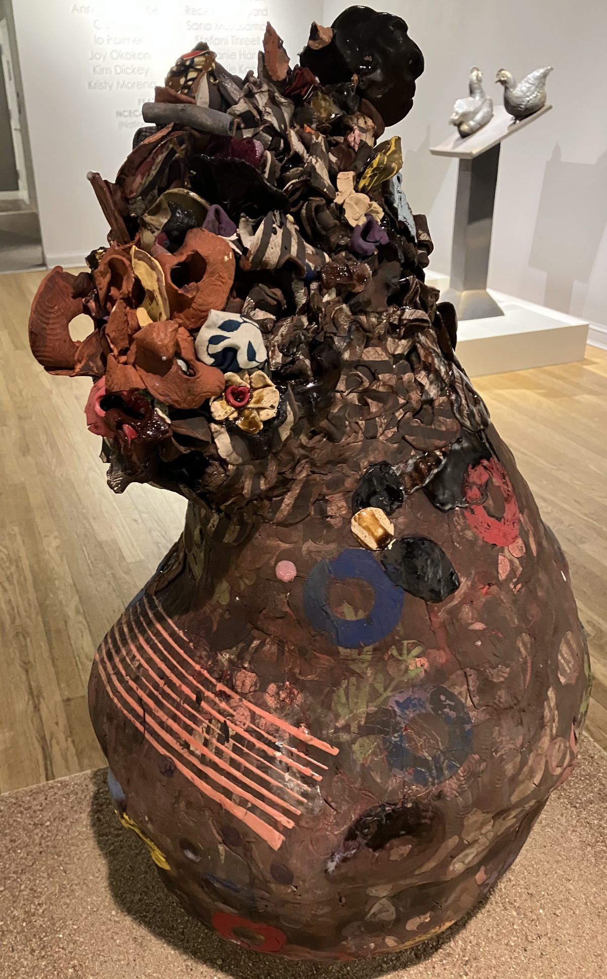 A large, textured ceramic sculpture resembling a distorted vessel. The surface is covered in colorful, organic forms and abstract patterns, with embedded objects and layers of glaze creating a richly detailed finish.