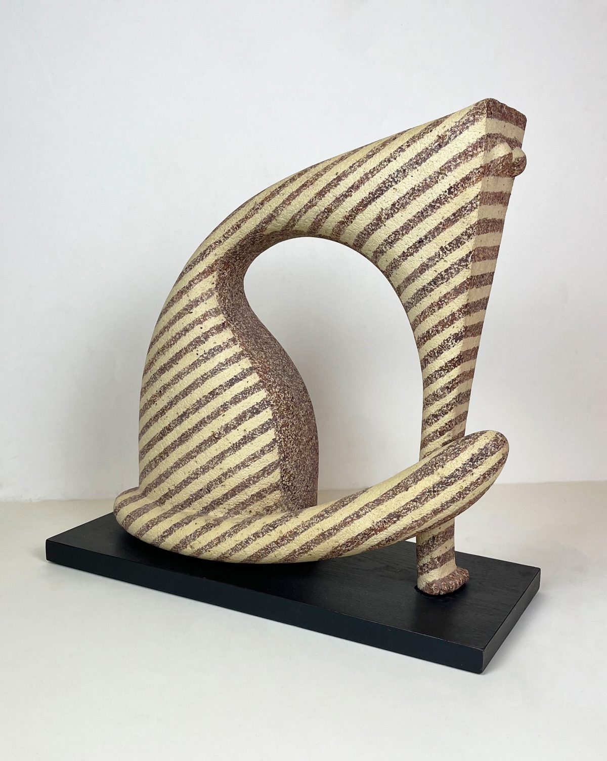 A ceramic sculpture featuring a swirling abstract form with brown diagonal stripes on a cream-colored surface, displayed on a black platform.