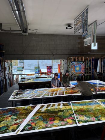 Three artists work in a stained glass studio, assembling colorful glass panels laid out across large tables.