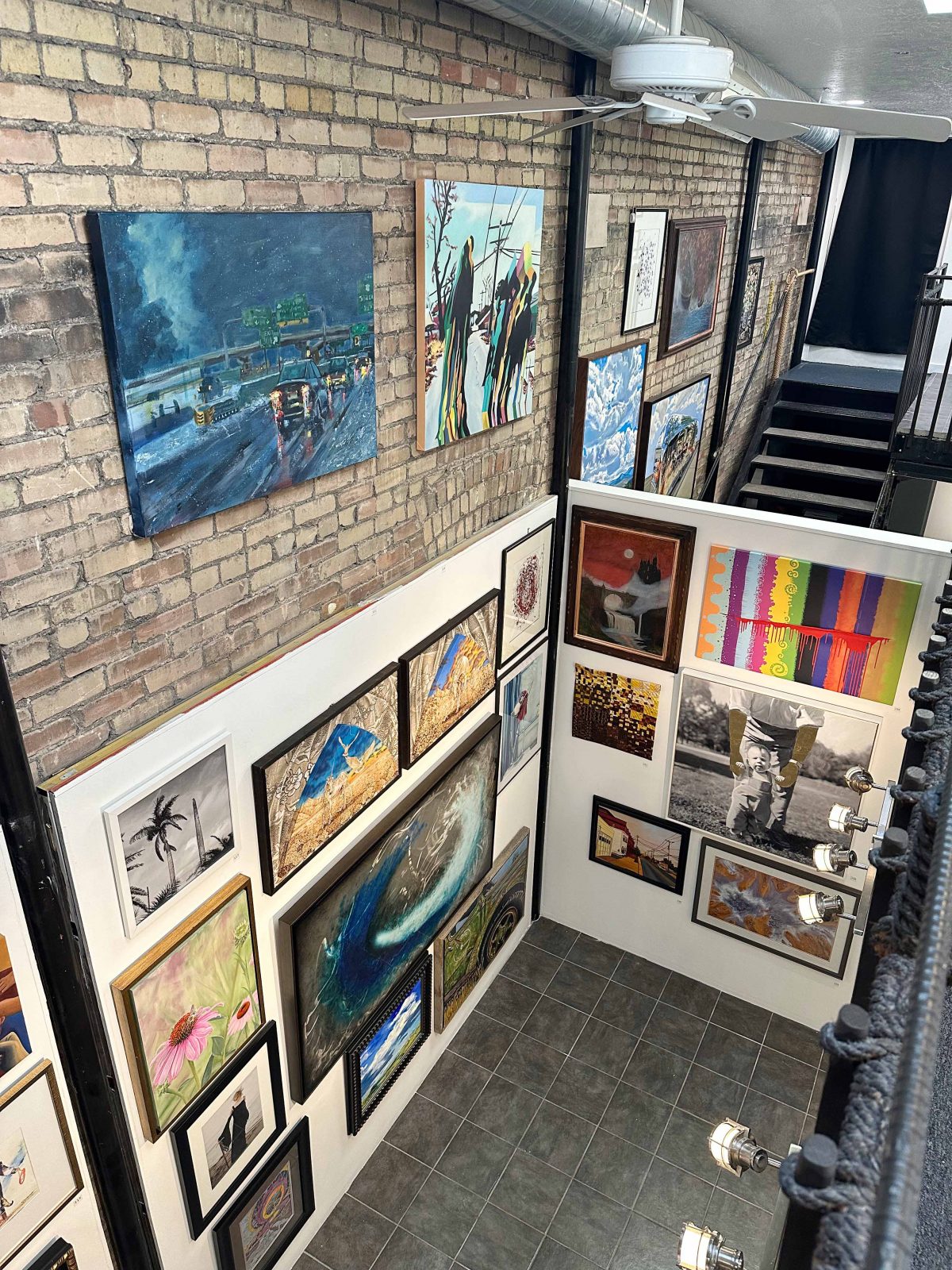 A brick-walled gallery space filled with colorful paintings and framed artworks, including a large tiger portrait, an expressive bear painting, and various abstract and landscape pieces arranged in a tight salon-style layout.